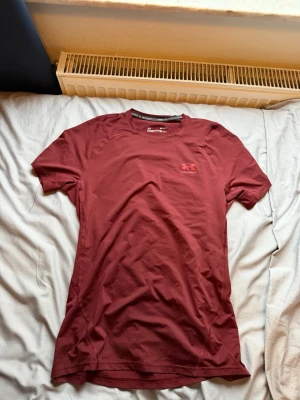 Vinröd tränings-tshirt Under Armour - Snygg vinröd tränings-tshirt från Under Armour med rund hals och korta ärmar. Tillverkad i funktionsmaterialet HeatGear som andas och transporterar bort fukt. Perfekt för gymmet eller löprundan. Diskret logga på bröstet.