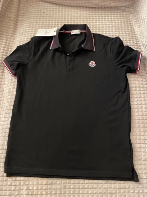 Svart pikétröja från Moncler - Klassisk svart pikétröja från Moncler med vit och röd detalj på krage och ärmslut. Tröjan har korta ärmar, knappslå framtill och Moncler-logga på bröstet. Tillverkad i mjuk bomull som känns skön mot huden. Perfekt för en clean och stilren look.