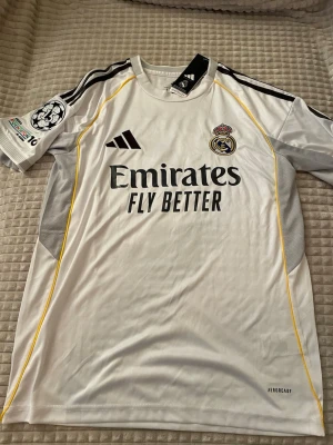 Real Madrid matchtröja Mbappé 10 - Snygg vit Real Madrid matchtröja från Adidas med Mbappé och nummer 10 på ryggen. Tröjan har svarta detaljer, gula linjer längs sidorna och klubbmärke samt sponsortryck på bröstet. Tillverkad i lätt och ventilerande material (AEROREADY). Perfekt för fotbollsträning eller match.