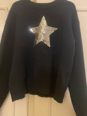 Svart stickad tröja med paljettstjärna - Cool marinblå stickad tröja med en stor stjärna i silvriga  paljetter på bröstet. Tröjan har rund halsringning och långa ärmar. Perfekt för dig som vill sticka ut lite extra med en glittrig detalj.