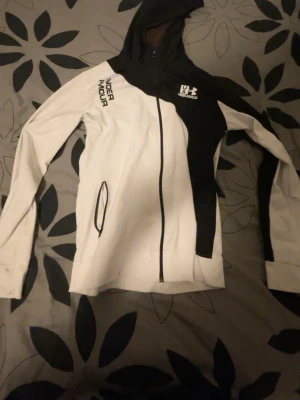 Svartvit hoodie från Under Armour - Snygg svartvit hoodie från Under Armour med dragkedja och huva. Stora loggor på bröstet och axeln, samt praktiska fickor med dragkedja. Sportig design i mjukt material, perfekt för en aktiv stil.