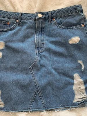 Blå jeanskjol med slitningar - Säljer en blå jeanskjol i kort modell med slitna detaljer och rå kant nertill. Kjolen har klassiska fem fickor och knappgylf. Perfekt för dig som gillar en avslappnad och trendig stil.