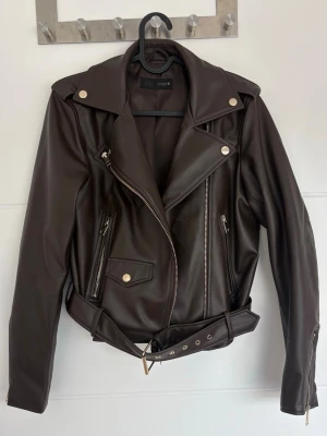 Brun bikerjacka från ZARA – stl S -   Snygg brun skinnjacka (fuskskinn) från ZARA i storlek S. Klassisk biker-modell med guldfärgade detaljer, sned dragkedja och flera praktiska fickor med dragkedjor. Justerbart midjebälte och bred krage som ger en riktigt stilren look.  Jackan är i fint skick och använd sparsamt. Inga skador, endast normalt bruksslitage. Tillverkad i Kambodja.  Perfekt för både vardag och fest – lätt att styla och sitter snyggt!
