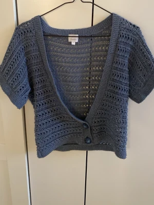 Blå stickad kortärmad kofta Camaieu - Snygg blå stickad kofta från Camaieu med korta ärmar och två knappar framtill. Koftan har ett luftigt hålmönster och djup v-ringning, perfekt att slänga över ett linne. Materialet känns mjukt och lätt, vilket gör den skön att bära under varmare dagar. Passar mig som är storlek xs-S 