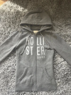 Grå zip hoodie från Hollister - Säljer en grå hoodie från Hollister med dragkedja och vit text på bröstet. Hoodien har två fickor framtill, justerbar huva med snören och mjukt, fleecefodrat material. Perfekt för chill dagar eller när du vill ha en avslappnad look. Lite nopprig