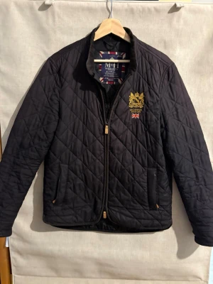 Svart quiltad jacka från Morris - Snygg svart quiltad jacka från Morris med guldbroderad logga och brittisk flagga på bröstet. Jackan har dragkedja framtill, ståkrage och två fickor. Perfekt för dig som gillar klassisk stil med en modern twist.