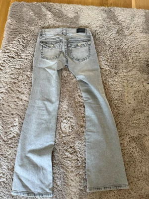 Ljusgrå bootcut jeans från Gina Tricot  - Säljer ett par ljusgrå bootcut jeans från gina tricot med klassiska fem fickor och snygga detaljer på bakfickorna. Jeansen har låg midja och är tillverkade i ett mjukt denimtyg som sitter skönt. Perfekta för dig som gillar en avslappnad men trendig look. Säljs ej längre i butik