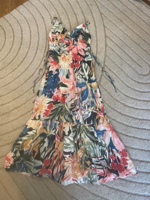 Populär slutsåld Blommig långklänning med snörning - Säljer en populär slutsåld  färgglad långklänning med blommigt mönster i rosa, blått, grönt, gult och rött. Klänningen har smala axelband, v-ringning och snörning i midjan för en snygg passform. Perfekt för sommarens alla tillfällen!