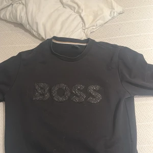 Svart sweatshirt från BOSS - Snygg svart t-shirt från BOSS med stor logga i svart på bröstet. Klassisk rund halsringning och långa ärmar. Perfekt för dig som gillar stilrena plagg med tydlig branding.