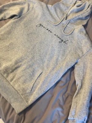 Grå hoodie med texttryck - Snygg grå hoodie med huva och dragsko. På bröstet står det 'you are enough' i svart skrivstil. Hoodien har en stor magficka och breda muddar vid ärmar och nederkant. Perfekt för en chill och avslappnad stil.