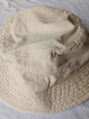 Beige bucket hat i bomull - Säljer en stilren beige bucket hat i mjuk bomull. Hatten har klassisk form med rundad kulle och bred, nedåtlutande brätte. Perfekt för en avslappnad streetwear-look och enkel att matcha med olika outfits.