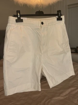 Vita shorts med resårmidja - Säljer ett par stilrena vita shorts med resår i midjan och knappstängning. Shortsen har raka ben och sidofickor, perfekta för varma dagar. Materialet känns lätt och luftigt, vilket gör dem sköna att bära hela sommaren.