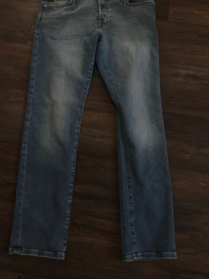 Blå raka jeans från Jack & Jones - Säljer ett par klassiska blå jeans från Jack & Jones med rak passform. Jeansen har fem fickor, bälteshällor och snyggt slitna detaljer framtill. Perfekta för en avslappnad och trendig look. Materialet är mjukt denim i bomull.  Dem har ej använts helt nya. 
