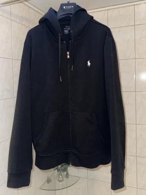 Polo Ralph Lauren Zip Hoodie - Snygg svart hoodie med dragkedja från Polo Ralph Lauren. Klassisk design med huva och dragsnören, samt fickor framtill. Ikoniska vita loggan broderad på bröstet. Perfekt för en avslappnad och stilren look. Färg: svart. Självklart äkta. Mycket bra skick! Nypris: 2229kr. Tar endast Swish. Spårbar och säker frakt med PostNord funkar även fint. Kör ej här via Plick, kör utanför denna appen!