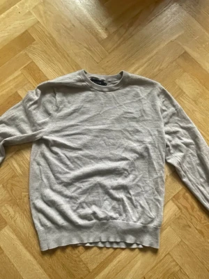 Ljusgrå stickad tröja från Riley, stickad tröja, knitt - En stilren ljusgrå stickad tröja från Riley med rund halsringning och långa ärmar. Tröjan har ribbade muddar vid ärmslut och nederkant, vilket ger en klassisk och enkel look. Perfekt att matcha med jeans eller chinos.