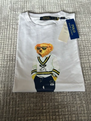 Vit Polo Bear t-shirt från Ralph Lauren - Säljer en vit t-shirt från Ralph Lauren med den ikoniska Polo Bear-trycket på framsidan. T-shirten har rund hals, korta ärmar och är gjord i mjuk bomull. Perfekt för dig som gillar klassisk och stilren streetwear med en twist.