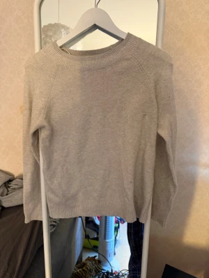 Beige stickad tröja med rund hals - En enkel och stilren beige stickad tröja med rund halsringning och långa ärmar. Tröjan har ribbade muddar vid ärmslut och nederkant. Perfekt för lager på lager och passar till det mesta i garderoben. Har ett litet hål så pris går att diskutera