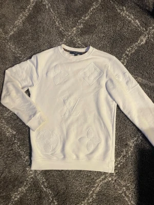 Vit LV tröja  - Säljer en clean vit sweatshirt från Louis Vuitton i storlek M. Tröjan har broderade LV-loggor och monogramdetaljer ton-i-ton framtill. Klassisk rund halsringning och långa ärmar. Perfekt för en clean look.