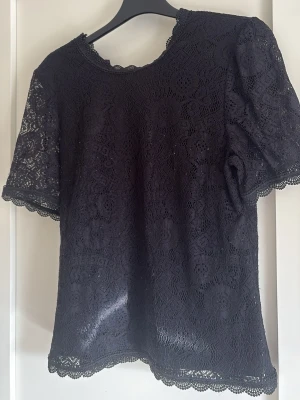 Svart spetsblus med kort ärm - Svart blus i spets med korta ärmar och rund halsringning. Blusen har fina spetsdetaljer och vågig kant vid ärmslut och nederkant. Perfekt för dig som gillar en romantisk och stilren look.
