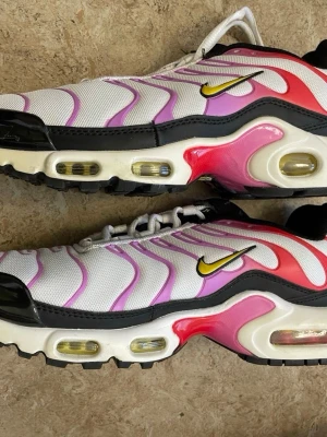 Nike Air Max Plus vita/rosa sneakers - Nike Air Max Plus sneakers i vitt mesh med rosa och lila detaljer, svarta och röda inslag samt gula Swoosh-loggor. Synlig Air Max-dämpning i sulan och svarta gummipartier. Ikonisk vågig design och robust yttersula för extra grepp.