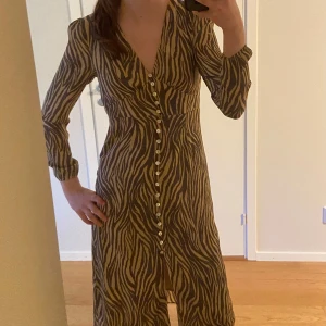 Zebramönstrad långklänning Adoore - Snygg långklänning från Adoore  med zebramönster i beige och svart. Klänningen har v-ringning, långa ärmar och knappar framtill. Perfekt för dig som vill sticka ut med ett trendigt djurmönster.