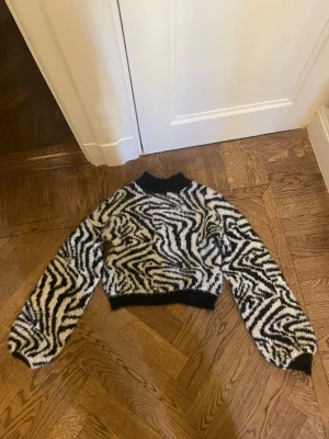 Zebramönstrad fluffig tröja - Säljer en supermjuk och fluffig zebramönstrad tröja med svartvitt mönster. Tröjan har svarta muddar vid hals, ärmslut och midja samt en croppad passform. Perfekt för dig som vill sticka ut med en trendig look.