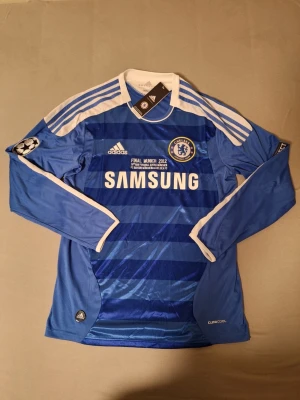 Chelsea långärmad fotbollströja 2011/2012 - Chelsea FC långärmad matchtröja från Adidas i blått med vita detaljer och tre ränder på ärmarna. Tryck med Samsung-logga, Champions League-märke och FINAL MÜNCHEN 2012 på bröstet. Denna tröja är från den ikoniska Champions League-finalen 2012 där Chelsea mötte Bayern München. En historisk match där Chelsea tog sin första Champions League-titel efter en dramatisk final som avgjordes på straffar.Skicka gärna meddelande vid frågor. Snabb affär = snabb leverans 📦 Skickas oftast samma eller nästa dag