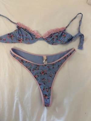 Blommig bikini  - Sött bikini med blå botten och rosa blommönster. Bh:n har tunna axelband och rosa volangdetaljer längs kuporna. Matchande trosa med hög skärning och rosa kantband. Materialet känns mjukt och stretchigt, perfekt för en feminin look.   Aldrig använd har dock klippt av lappen, Bh:n är i storlek 70 A motsvarande XS.