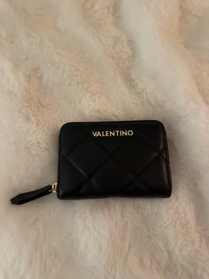 Svart quiltad plånbok från Valentino - Snygg svart plånbok från Valentino med quiltat mönster och guldfärgad logga framtill. Plånboken har dragkedja runtom och flera fack för kort och mynt. Tillverkad i konstläder med stilren design och praktisk storlek.