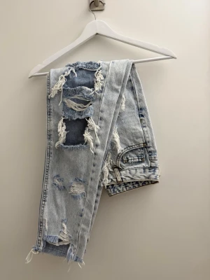 Ljusblå ripped mom jeans - Säljer ett par ljusblå mom jeans med kraftiga slitningar och stora hål framtill. Jeansen har hög midja, raka ben och råa kanter vid bensluten. Perfekta för en trendig och avslappnad streetstyle-look.