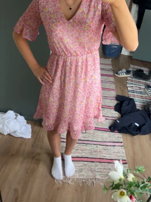 Rosa blommig kortklänning med volang - Söt rosa klänning med blommigt mönster och volangdetaljer vid ärmar och nederkant. Klänningen har v-ringning, resår i midjan och korta ärmar.