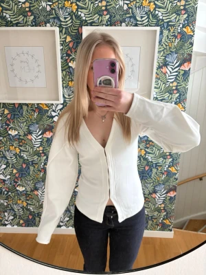 Vit kofta från Nelly - Snygg vit kofta med v-ringning som man knäpper framtill. Koftan har långa ärmar och är tillverkad i ett mjukt material som ger en clean och stilren look. 