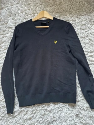 Svart stickad tröja från Lyle & Scott - Klassisk svart stickad tröja från Lyle & Scott med V-ringning och den ikoniska gula örnloggan broderad på bröstet. Tröjan har långa ärmar och ribbade muddar vid ärmslut och nederkant. Perfekt för lager-på-lager och enkel att matcha.