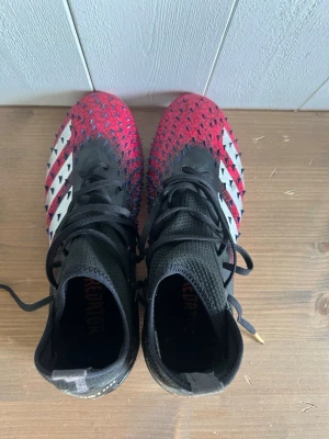 Adidas Predator fotbollsskor röd/svart - Säljer ett par Adidas Predator fotbollsskor i rött och svart med vita detaljer och klassiska tre ränder på sidan. Skorna har hög krage i stickat material för extra stöd och snörning för bra passform. Ovandelen har gummidetaljer för bättre bollkontroll.