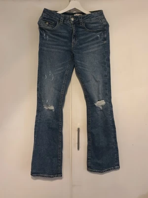 Blå bootcut jeans med slitningar - Snygga blå bootcut jeans med slitna detaljer på knäna och lätt tvättad look. Klassisk femficksmodell med knapp och dragkedja framtill. Perfekta för en avslappnad och trendig stil.