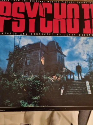 Psycho II original motion picture soundtrack LP-skiva - Soundtracket är komponerat och dirigerat av Jerry Goldsmith till filmen Psycho II. Formatet är vinyl LP med omslag som visar filmens husmotiv på framsidan och filmrelaterad information på baksidan. Skivan är avsedd för uppspelning på skivspelare med standard 33 1/3 rpm-inställning. Omslaget har synligt slitage och en skada i ett hörn, enligt bilderna. Inga repor.