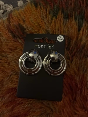 Silverfärgade ringörhängen Montini - Säljer ett par stilrena örhängen från Montini i silverfärgad metall. Örhängena har en modern design med dubbla cirklar som ger en cool och trendig look. Perfekta för att lyfta vilken outfit som helst med en touch av metallic.