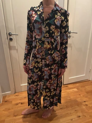 Blommig långklänning med krage - Snygg långklänning med färgglatt blommönster i svart, blått, gult och rött. Klänningen har lång ärm, knappar framtill och klassisk krage. Perfekt för dig som gillar statement-plagg med mycket färg och mönster.