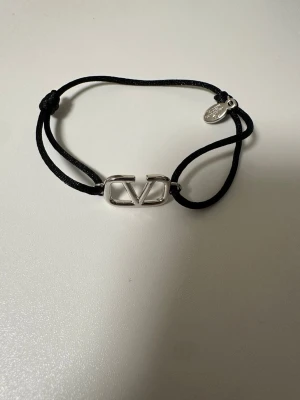 Valentino svart armband med logga - Snyggt svart armband från Valentino med justerbart band i textil och en silverfärgad metallogo i mitten. Armbandet har även en liten rund bricka med ingraverad text. Perfekt accessoar för att lyfta din stil.