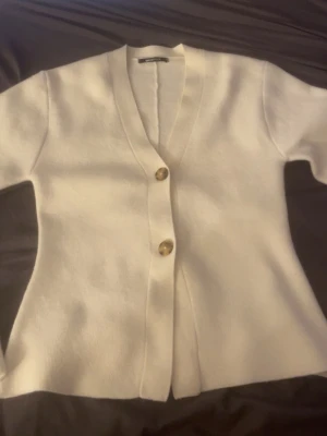 Vit kofta med träknappar - Stilren vit kofta med två dekorativa träknappar framtill och djup V-ringning. Koftan har lång ärm och är tillverkad i ett mjukt, stickat material som ger en avslappnad men ändå chic look. Perfekt att slänga över en topp för extra lager.