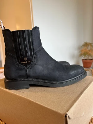 Svarta boots i mocka från Gant - Snygga svarta stövletter från Gant i mjuk mocka med elastiska paneler på sidorna och dragflikar med logga baktill. Klassisk rund tå och låg klack gör dem perfekta till många olika stilar. Tidlös design med diskret Gant-märke på sidan.