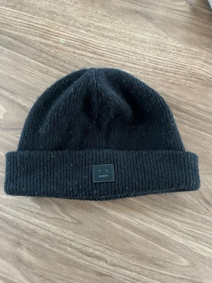 Svart ribbstickad mössa Acne Studios - Säljer en svart ribbstickad mössa från Acne Studios med uppvikt kant och den klassiska Face-loggan framtill. Mössan är gjord i mjukt material och har en enkel, stilren design som passar till det mesta. Modellen heter plain Beanie och är köpt på Acne Studios i Stockholm för 1600 kr. Skick 9/10 och använd 3-4 gånger endast 