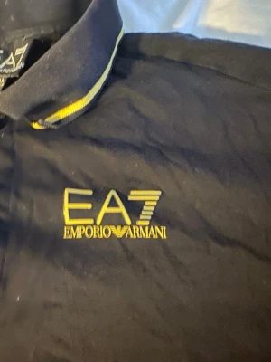Svart EA7 Emporio Armani piké - Snygg svart pikétröja från EA7 Emporio Armani med gul logga broderad på bröstet. Kragen har gula detaljer som ger en sportig touch. Perfekt för dig som vill ha en stilren och trendig look. Tröjan är för barn. Tröjan har storleken 160 men är lite liten i storleken 