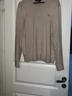 Beige Morris tröja - Stilren beige tröja med rund hals och diskret broderad logga på bröstet. Tröjan har långa ärmar och ribbade muddar vid ärmslut och nederkant. Perfekt att matcha med jeans för en clean look.