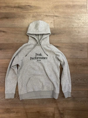 Peak performance hoodie  - Ljusgrå hoodie från Peak Performance med svart logotyp och text 'Est. 1986' på bröstet. Klassisk känguruficka framtill, ribbade muddar och dragsko i huvan. Tillverkad i mjukt bomullsmaterial som känns skönt mot huden. Perfekt för en avslappnad och sportig stil. Priset går att diskutera.