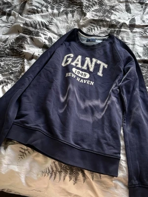 Mörkblå sweatshirt från GANT - Säljer en mörkblå sweatshirt från GANT med tryck 'GANT 1949 NEW HAVEN' på bröstet. Tröjan har rund halsringning, långa ärmar och ribbade muddar. Perfekt för en avslappnad och sportig stil.