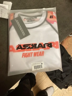 Askari rashguard och fight shorts - Säljer ett set med rashguard och fight shorts från Askari Fight Wear. Tröjan är vit med röda och svarta detaljer, kort ärm och tight passform. Shortsen är svarta med vit och röd logga, elastisk midja och sportig design. Perfekt för MMA, grappling eller annan kampsport.