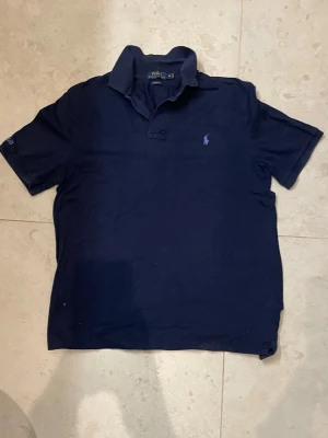 Mörkblå piké från Polo Ralph Lauren - Klassisk mörkblå pikétröja från Polo Ralph Lauren i bomull. Tröjan har korta ärmar, krage och knappar vid halsen samt den ikoniska broderade loggan på bröstet. Classic fit-modell som passar till det mesta.