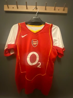 Arsenal Nike matchtröja Henry 14 - Säljer en klassisk Arsenal matchtröja från Nike med O2 sponsortryck och Henry 14 på ryggen. Tröjan är röd med vita ärmar och gula detaljer, har klubbmärke på bröstet och är tillverkad i lätt polyester. Perfekt för dig som älskar fotboll och Arsenal!