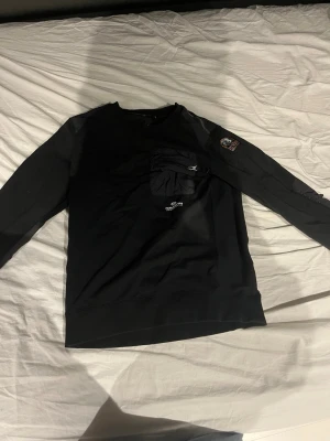 Svart sweatshirt från parajumper  - Cool svart sweatshirt från parajumper med rund halsringning och en unik ficka med dragkedja på bröstet. Patch med logga på ena ärmen och diskret tryck på bröstet. Perfekt för en avslappnad streetwear-look.